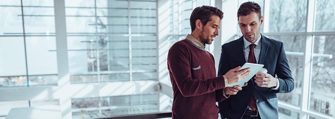 Two men in a modern office setting, one in a suit and the other in a casual sweater, looking at a tablet together, with large windows in the background letting in natural light